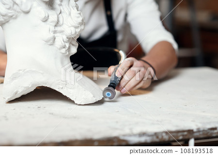 Professional bearded carver polishing with micro drill machine details of the man's sculpture head in his workshop. 108193518