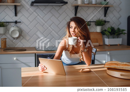 Pretty young woman using tablet while drinking morning coffee after breakfast on the kitchen. Pretty young woman using tablet while drinking morning coffee after breakfast on the kitchen. 108193585
