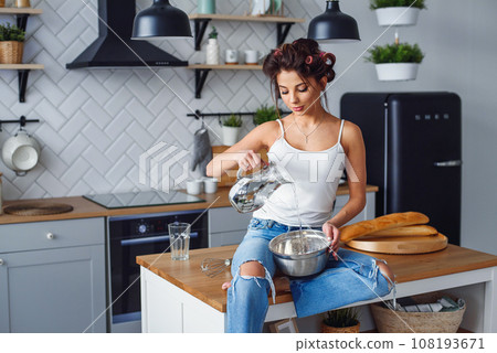 Beautiful smiling woman pours water in a deep metal plate to kneads a dough while sitting on the table in the modern stylish kitchen. Beautiful smiling woman pours water in a deep metal plate to kneads a dough while sitting on the table in the modern stylish kitchen. 108193671