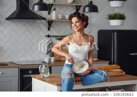 Beautiful smiling woman kneads a dough in a deep metal plate while sitting on the table in the modern stylish kitchen. Young pretty woman baking pie on the bright home kitchen. Beautiful smiling woman kneads a dough in a deep metal plate while sitting on the table in the modern stylish kitchen. Young pretty woman baking pie on the bright home kitchen. 108193681