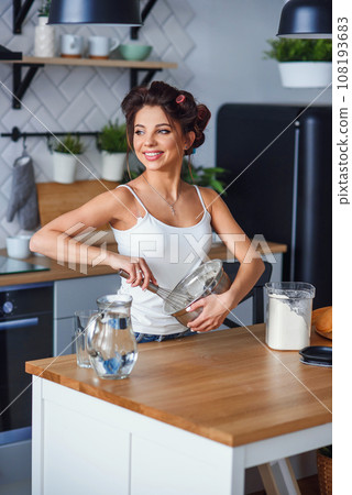 Beautiful smiling woman kneads a dough in a deep metal plate in the modern stylish kitchen. Young pretty woman baking pie on the bright home kitchen. 108193683