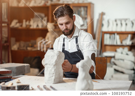 Male sculptor in white shirt and black apron makes a limestone copy of woman torso at the artistic studio. Male sculptor in white shirt and black apron makes a limestone copy of woman torso at the artistic studio. 108193752