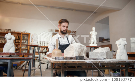 Male sculptor in white shirt and black apron makes a limestone copy of woman torso at the artistic studio. Male sculptor in white shirt and black apron makes a limestone copy of woman torso at the artistic studio. 108193777
