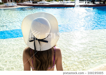 Back view of young woman with long hair wearing straw hat relaxing in summer near swimming pool with blue water on a sunny day. 108195005