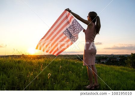 Back view of happy young woman posing with USA national flag outdoors at sunset. Positive female celebrating United States independence day. International day of democracy concept. 108195010