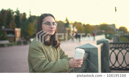 Young woman with a glass of coffee in her hand. A girl in glasses near the fence on the embankment with a hot drink in a glass. Young woman with a glass of coffee in her hand. A girl in glasses near the fence on the embankment with a hot drink in a glass. 108195108