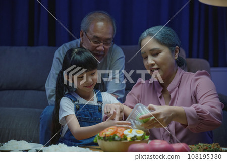 Happy Asian family grandmother grandfather and granddaughter dining on table and having fun during at home night time, senior parent and child eating food together in living room indoors together Happy Asian family grandmother grandfather and granddaughter dining on table and having fun during at home night time, senior parent and child eating food together in living room indoors together 108195380