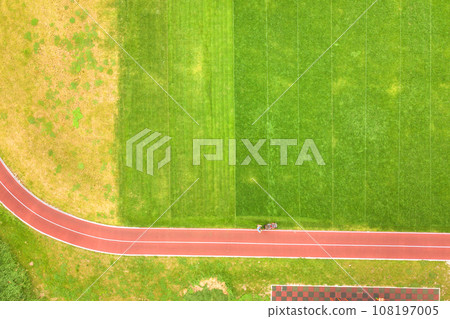 Aerial view of small figure of worker cutting green grass with mowing mashine on football stadium field with red running tracks in summer. 108197005