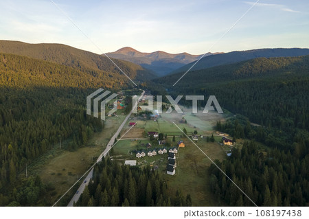 Aerial view of bright foggy morning over small rural homes between dark peaks with mountain forest trees at autumn sunrise. Beautiful scenery of wild woodland at dawn 108197438