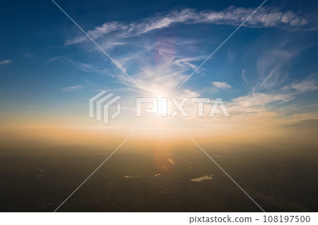 Aerial view from airplane window at high altitude of distant city covered with layer of thin misty smog and distant clouds in evening 108197500