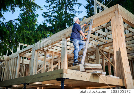 Carpenter constructing wooden frame house near the forest. Bearded man with glasses climbing ladder to the second floor of the two-story building. The concept of modern ecological construction. Carpenter constructing wooden frame house near the forest. Bearded man with glasses climbing ladder to the second floor of the two-story building. The concept of modern ecological construction. 108205172