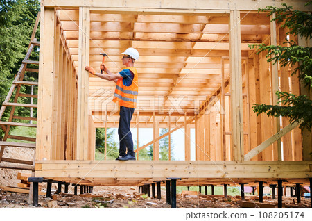 Wooden frame house being built by carpenter. Man wearing glasses hammering nails using hammer and is dressed in a protective helmet and a construction vest. Concept of modern ecological construction. Wooden frame house being built by carpenter. Man wearing glasses hammering nails using hammer and is dressed in a protective helmet and a construction vest. Concept of modern ecological construction. 108205174