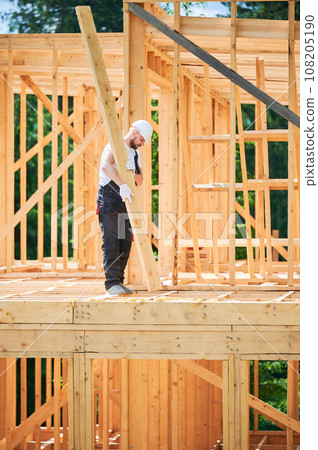 Carpenter constructs wooden-framed house. Man holds large beam in his hands while wearing work clothes and helmet. Idea of modern, eco-friendly construction. 108205190