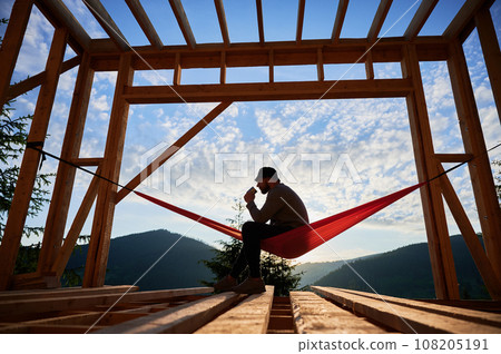 Man on construction site of wooden-framed house sits in hammock against backdrop of mountains and sunrise, sipping coffee. The concept pertains to modern ecological construction. Man on construction site of wooden-framed house sits in hammock against backdrop of mountains and sunrise, sipping coffee. The concept pertains to modern ecological construction. 108205191