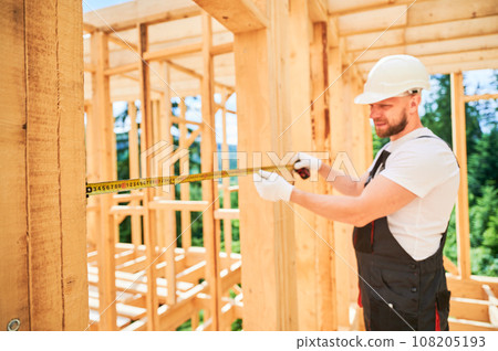 Carpenter building wooden frame house. Man measures the distance with tape measure while dressed in workwear and a helmet. Selective focus. Carpenter building wooden frame house. Man measures the distance with tape measure while dressed in workwear and a helmet. Selective focus. 108205193