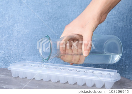 Woman pouring water into ice cube tray at light grey marble table, closeup Woman pouring water into ice cube tray at light grey marble table, closeup 108206945