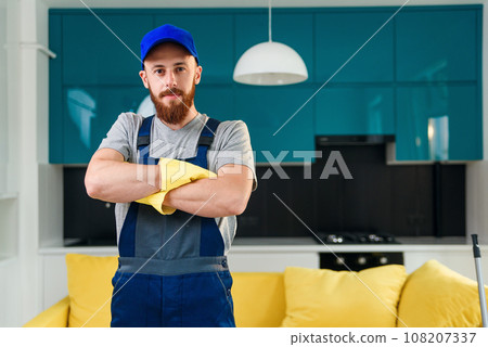 Handsome professional male worker from cleaning service in special uniform and yellow gloves folded his arms on the background of the contemporary cuisine. Handsome professional male worker from cleaning service in special uniform and yellow gloves folded his arms on the background of the contemporary cuisine. 108207337