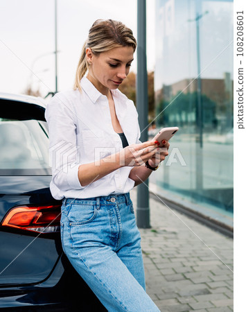 Woman looking at phone with serious face while leaning on car Woman looking at phone with serious face while leaning on car 108208601