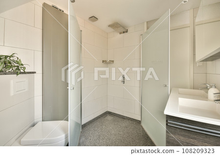 a bathroom with a toilet, sink and plant in the shower stall on the left side of the photo is an open white door 108209323