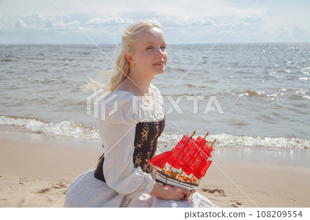 Portrait of romantic young blonde model woman in retro dress holding red sail boat on sea beach 108209554