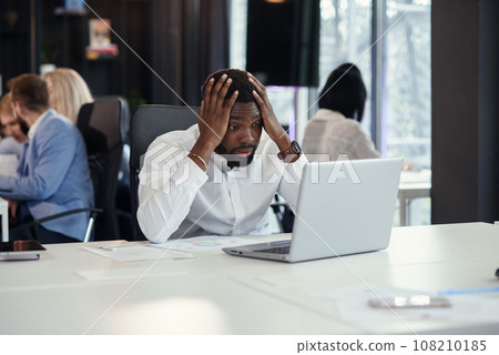 Disappointed african american office manager looking on the laptop screen and put his hands on head at his work table on the coworkers background. Disappointed african american office manager looking on the laptop screen and put his hands on head at his work table on the coworkers background. 108210185