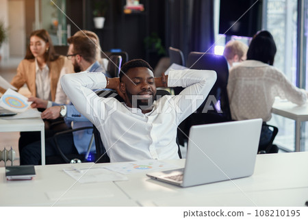Dark-skinned man with a little smile folded hands behind his head and looking at his laptop in modern office. 108210195