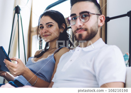 Couple using digital devices while sitting on bed and looking at camera 108210198