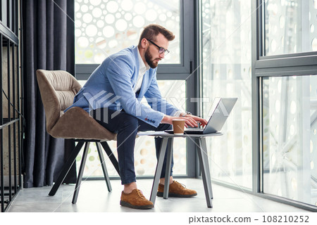 Handsome young male office manager with well-groomed beard and eyeglasses uses laptop and financial reports in the separate place in modern office centre. Handsome young male office manager with well-groomed beard and eyeglasses uses laptop and financial reports in the separate place in modern office centre. 108210252
