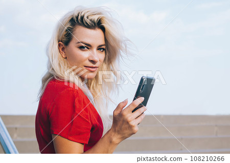 Confident woman with smartphone looking at camera on stairs Confident woman with smartphone looking at camera on stairs 108210266