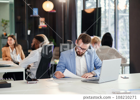 Successful bearded businessman in glasses and in blue suit works with laptop in modern office. 108210271