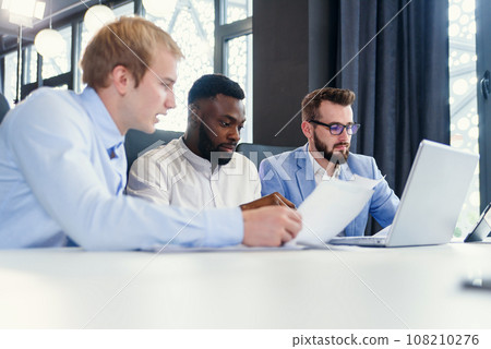 Three mixed races handsome businessmen discussing opportunities of their business strategy using different reports with infographics. Three mixed races handsome businessmen discussing opportunities of their business strategy using different reports with infographics. 108210276