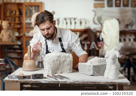 Stonemason which sitting in creative workshop and scraping the remains of lime-stone using brush 108210320
