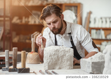 Handsome sculptor brushing stone head sculpture on the table in the atmospheric studio 108210329