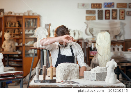 Male sculptor in white shirt and black apron makes a limestone copy of woman torso at the artistic studio. Male sculptor in white shirt and black apron makes a limestone copy of woman torso at the artistic studio. 108210345