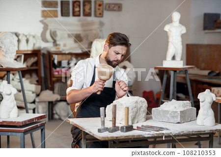 Bearded craftsman works in white stone carving with a chisel. Creative workshop with works of art. 108210353