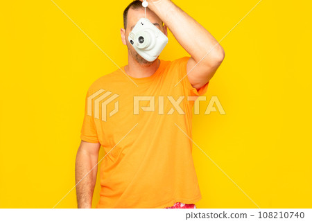 Bearded Hispanic man in his 40s holding an instant camera in front of his face, isolated on yellow studio background 108210740