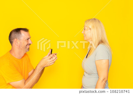 Man proposing to a woman who looks happy and surprised. Concept of love and marriage. isolated yellow background. Romantic concept, buying jewelry, wedding plans for bride and groom. 108210741