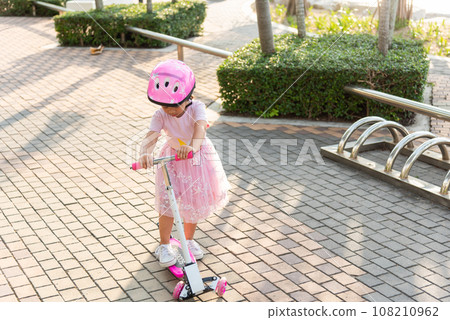 Child riding scooter. Happy Asian little kid girl wear safe helmet playing pink kick board on road in park outdoors on summer day, Active children games outside, Kids sport healthy lifestyle concept 108210962