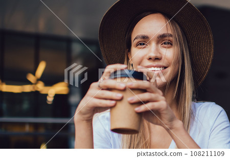 Joyful trendy woman drinking coffee while reading book in cafe 108211109