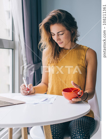 Focused female sitting at table with notebook and cup filling documents Focused female sitting at table with notebook and cup filling documents 108211361