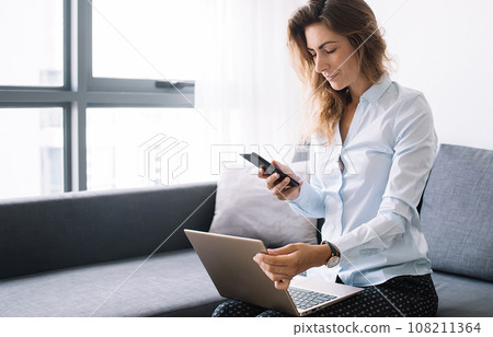 Lady using phone sitting with laptop on sofa 108211364