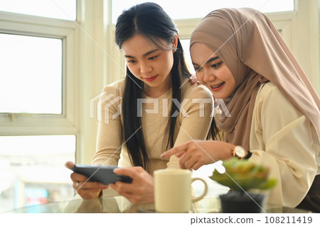 Smiling female friends playing game or watching video on mobile phone together during lunch break 108211419