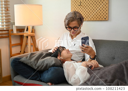 Romantic young couple in earphones watching social media videos on smartphone while sitting on couch Romantic young couple in earphones watching social media videos on smartphone while sitting on couch 108211423