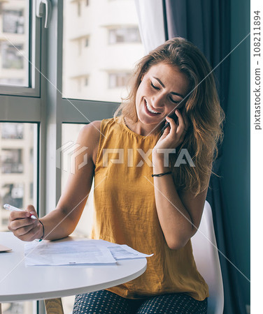 Stylish woman talking on smartphone and sitting at table with laptop and papers Stylish woman talking on smartphone and sitting at table with laptop and papers 108211894