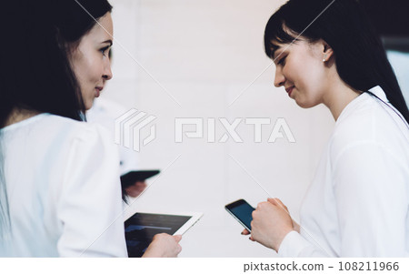 Female coworkers talking while using gadgets Female coworkers talking while using gadgets 108211966
