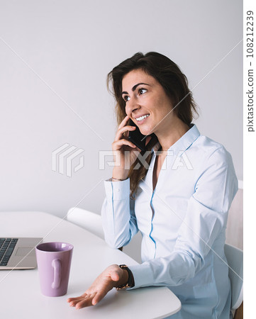 Woman in shirt talking on phone sitting with computer and coffee on table 108212239
