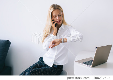 Attentive female sitting at table with laptop talking on mobile and looking at watch Attentive female sitting at table with laptop talking on mobile and looking at watch 108212408