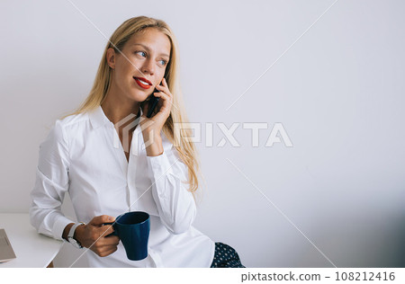Focused female sitting at table talking on phone and working on laptop 108212416