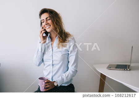 Smiling young brunette with cup sitting and talking on smartphone 108212617