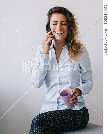 Smiling young woman with eyes closed and cup talking on smartphone 108213141